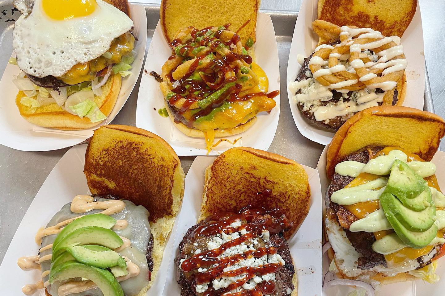 Six gourmet burgers rest on a tray, each topped with unique ingredients like avocado, fried egg, sauces, and cheese, held in toasted buns.