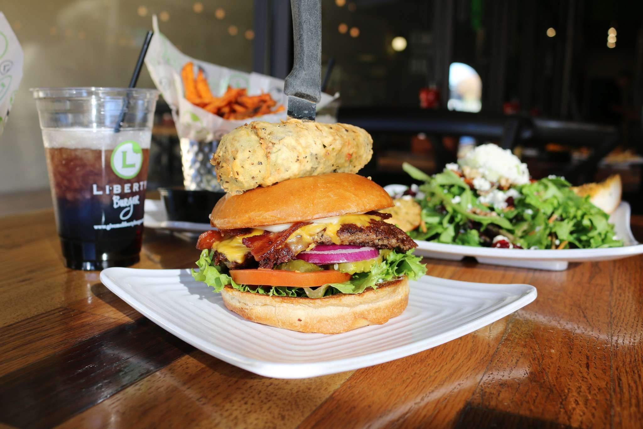 Burger with knife inserted, topped with biscuit, placed on white plate; salad, fries, and drink in background on wooden table. "LIBERTI" visible on cup.
