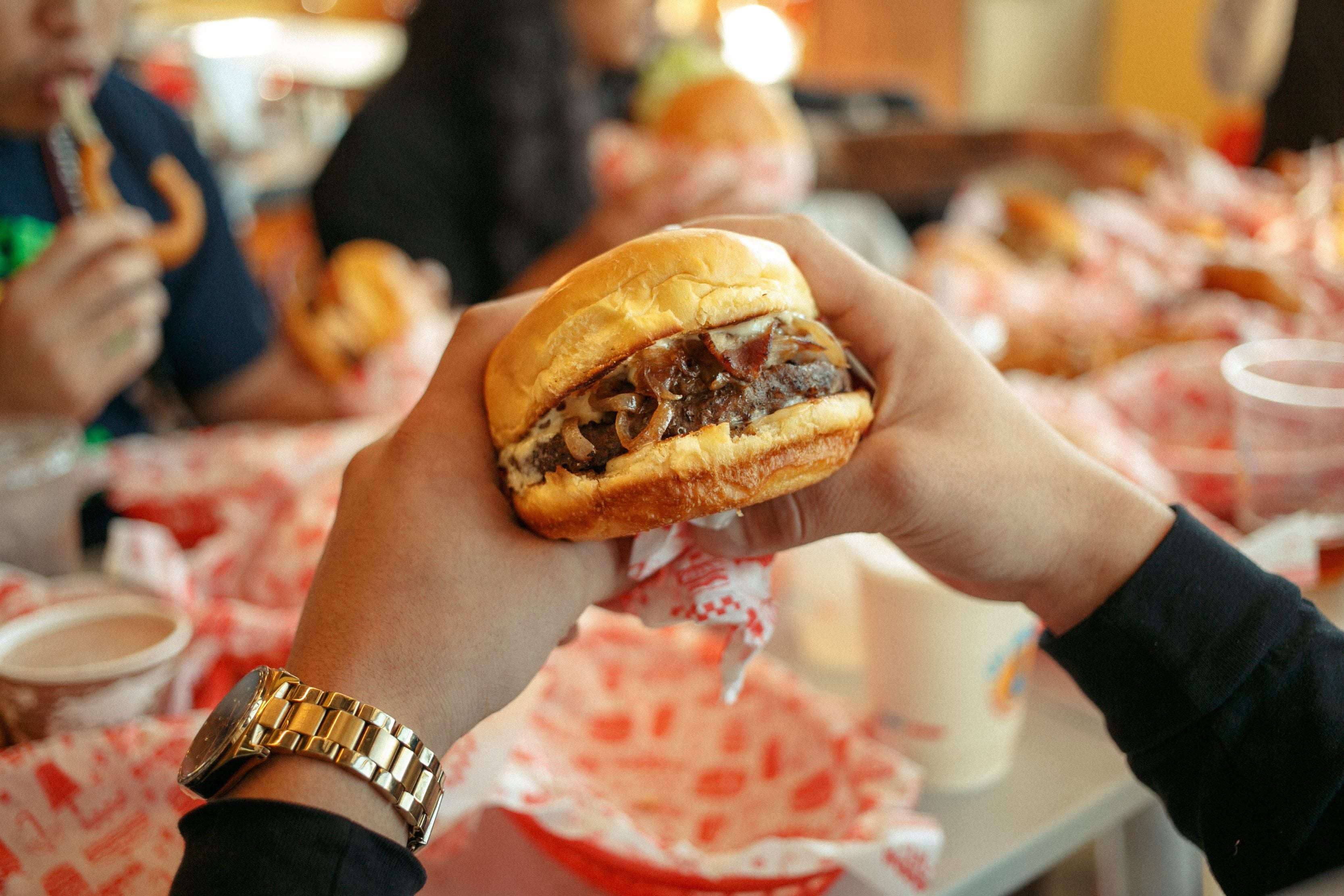 Burger held by hands with a gold watch, surrounded by people eating at a table with red-patterned wrappers and drinks.