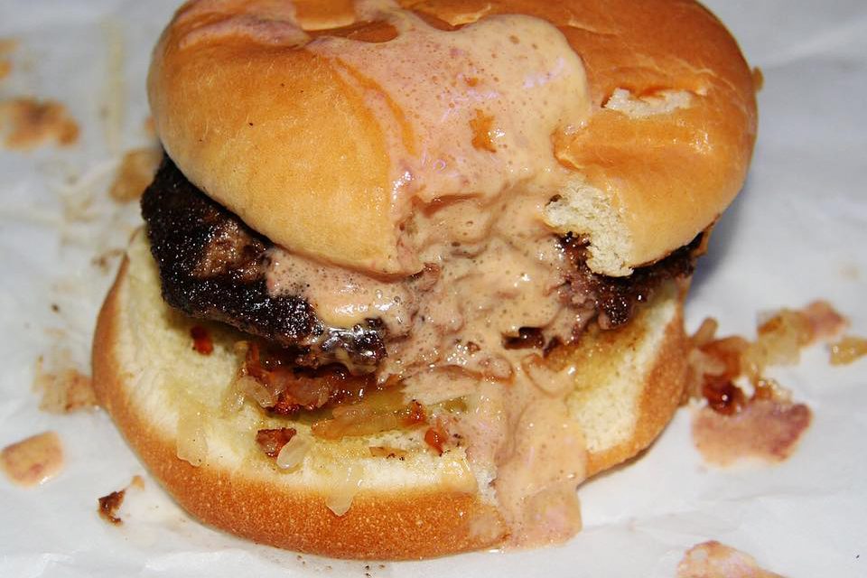 Burger oozing melted cheese and sauce, resting on a white paper surface with scattered crumbs and drips.