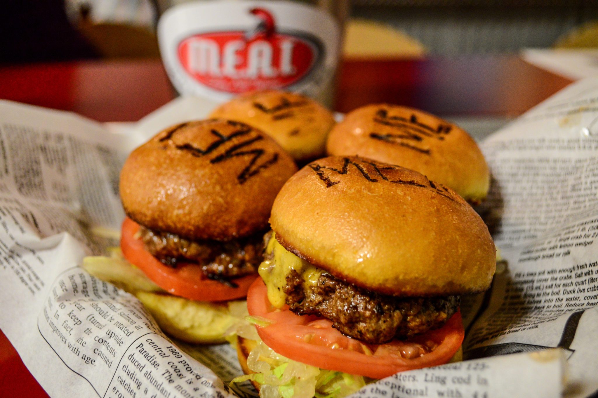 Burgers sit on newspaper-style paper in a restaurant. They have branded buns, visible toppings, and a blurred cup in the background with "MEAL" text.