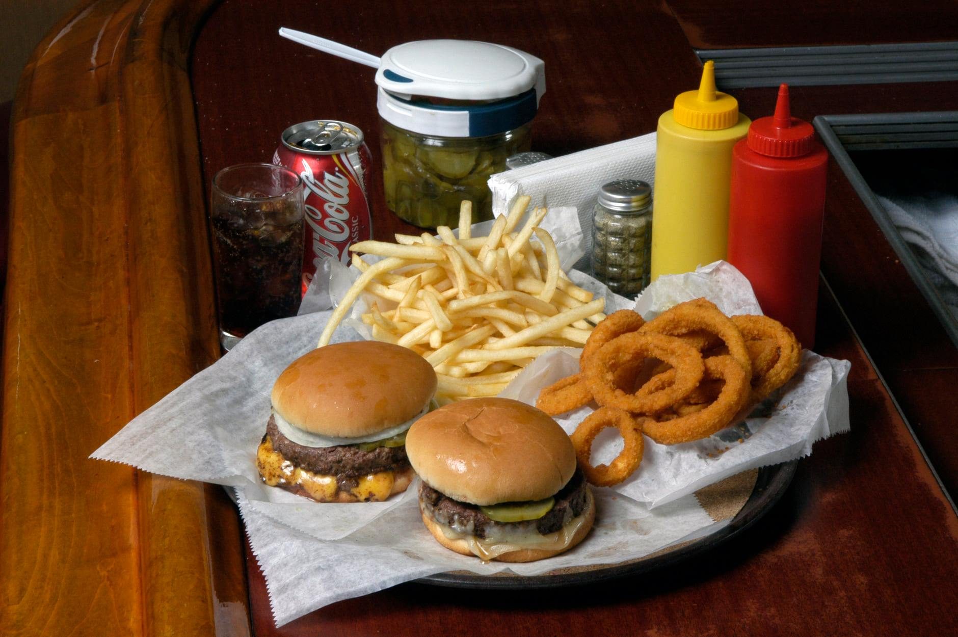 Burgers rest on a tray with fries and onion rings. Condiments, pickles, and a Coke can sit on a wooden table in the background.