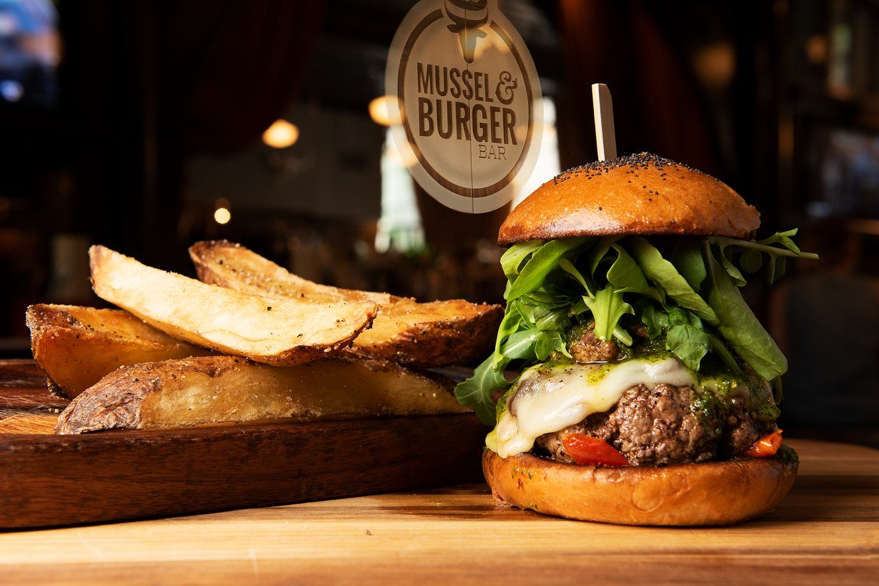Burger stacked with lettuce, cheese, and sauce beside large potato wedges on a wooden board in a restaurant setting. Sign reads "Mussel & Burger Bar."