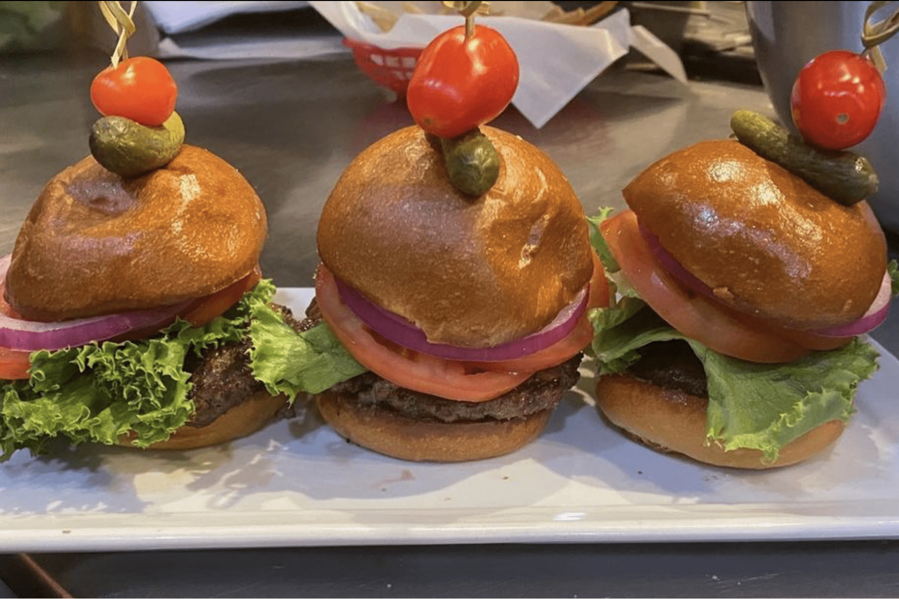 Three mini burgers displayed on a rectangular plate, each garnished with lettuce, tomato, onion, a pickle, and a cherry tomato skewer, in a kitchen setting.