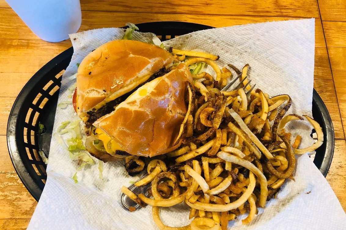 Burger and curly fries sit on a paper towel-lined basket, surrounded by a wooden table with a cup nearby.