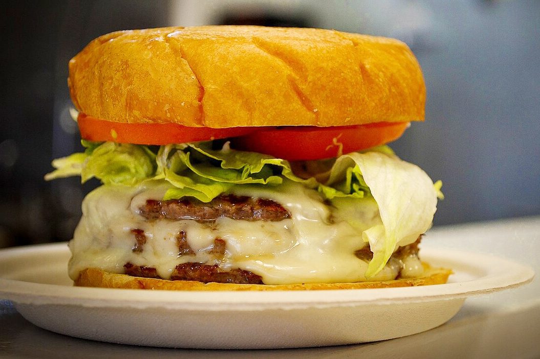 Burger sits on a plate with melted cheese, lettuce, and tomato inside a bun, against a blurred background.