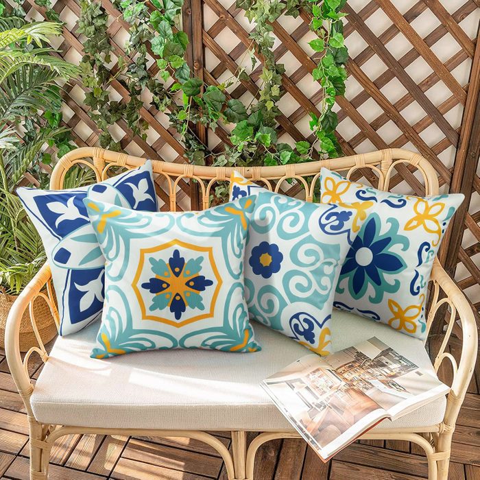 Colorful patterned cushions rest on a wicker bench, surrounded by leafy plants and a wooden trellis, with an open magazine nearby.