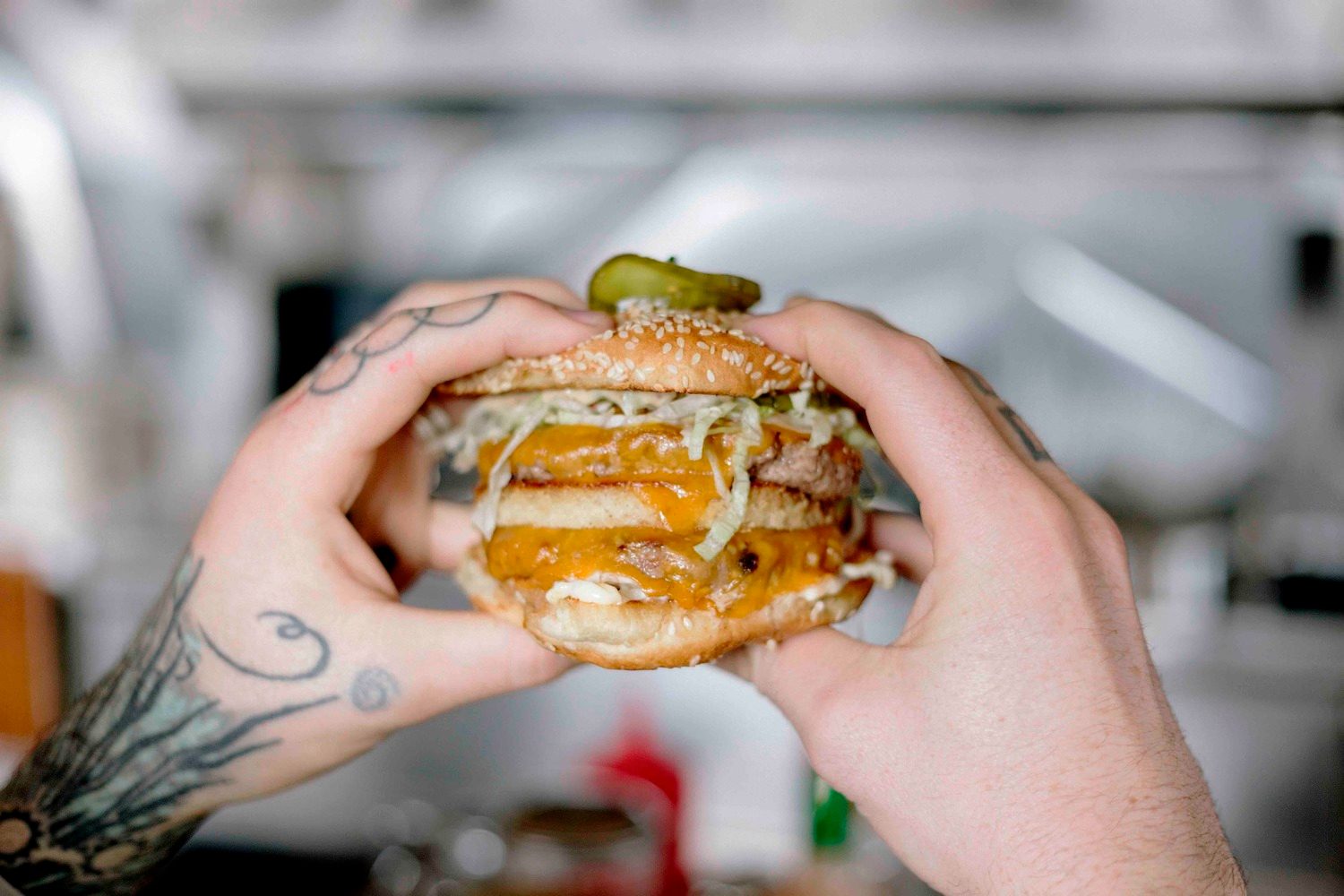 Tattooed hands hold a large burger with multiple layers, cheese, and lettuce in a blurred kitchen setting.