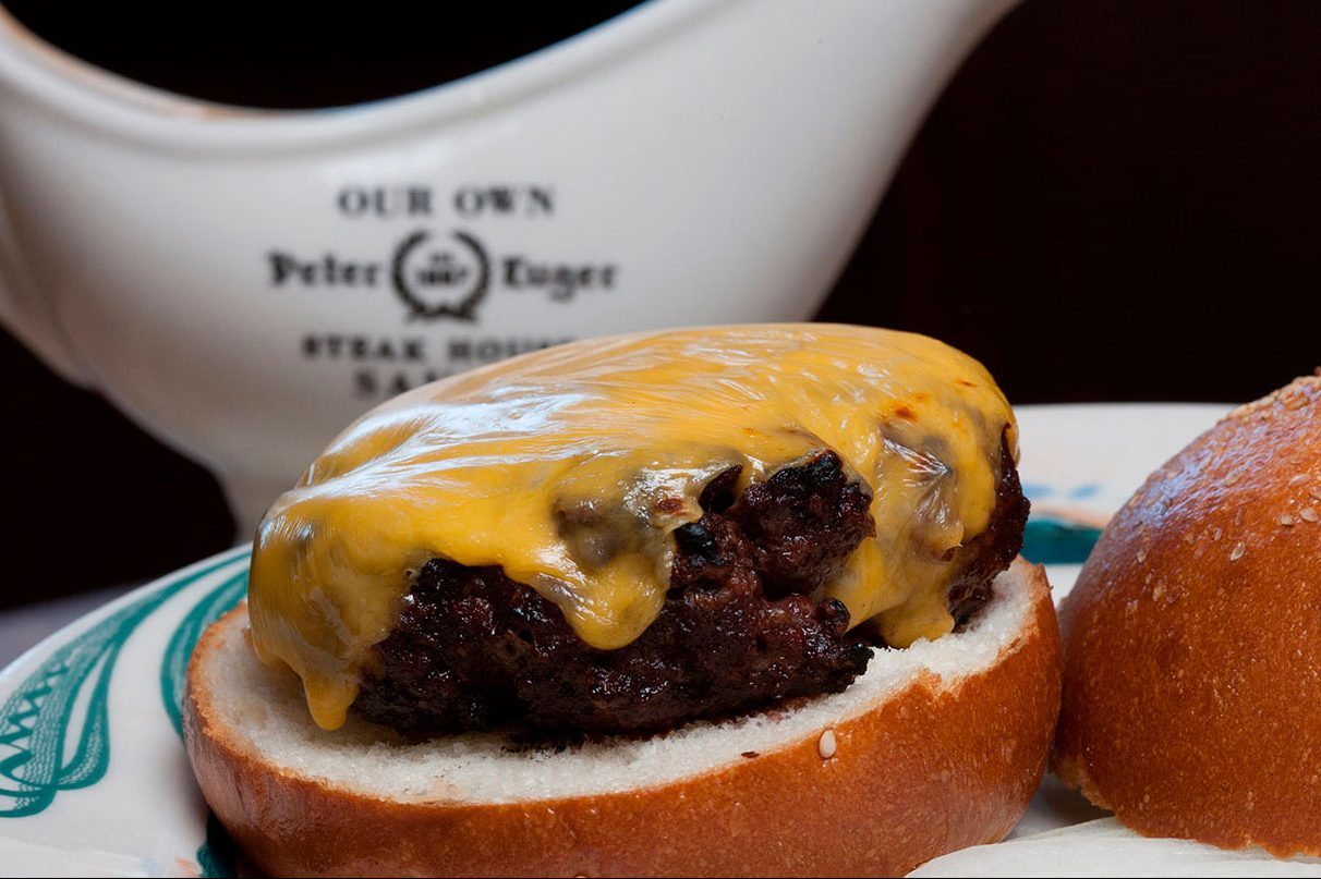 Cheeseburger sits on a bun, with cheese melting over the patty, placed on a plate near a sauce container labeled "OUR OWN Peter Luger STEAK HOUSE."