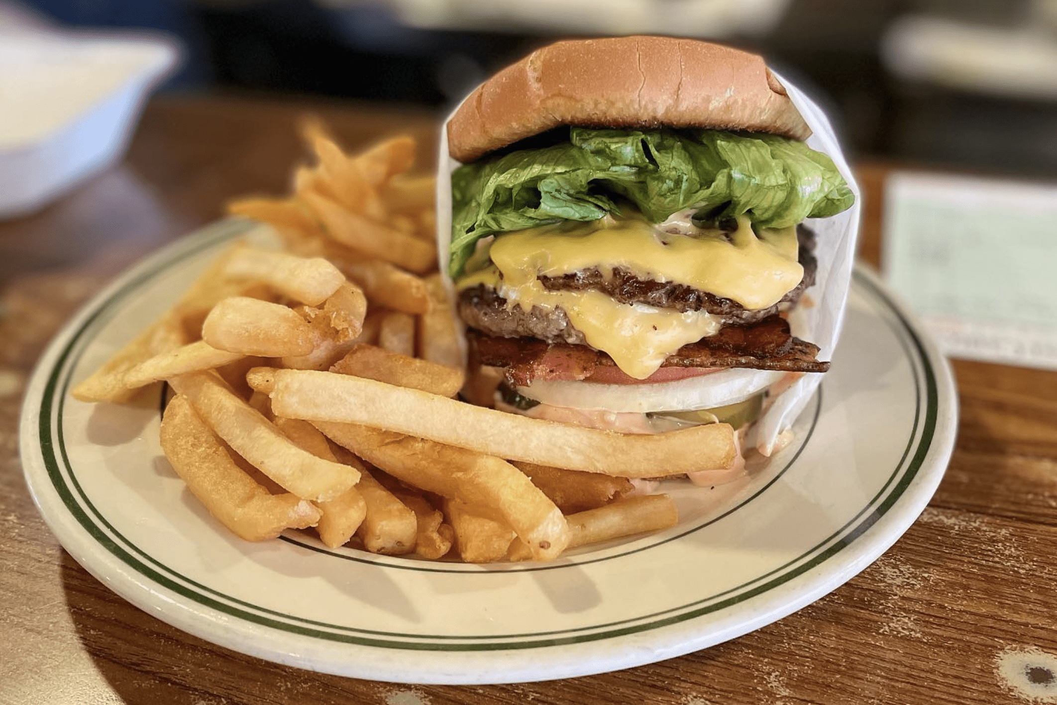 Burger sits on a plate beside fries, atop a wooden table; features cheese, lettuce, bacon, and a bun.