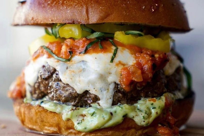 Burger stacked with melted cheese, chunky tomato salsa, and sliced pickles, set between a toasted bun, in a close-up food setting.
