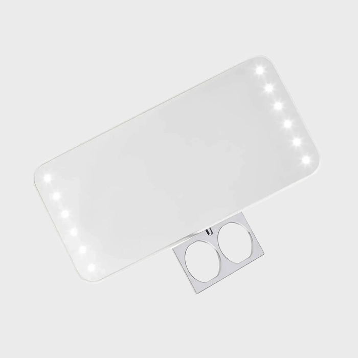 Rectangular lighted mirror emitting soft white light, positioned diagonally, featuring a folding metallic stand against a plain light gray background.