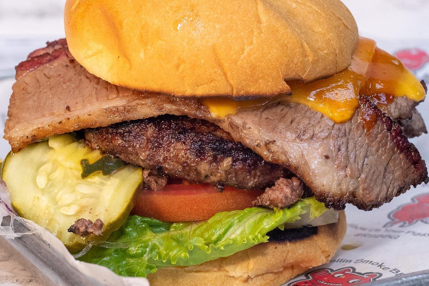 Burger topped with brisket and cheddar, resting on lettuce, tomato, and pickles in a bun, placed on paper.