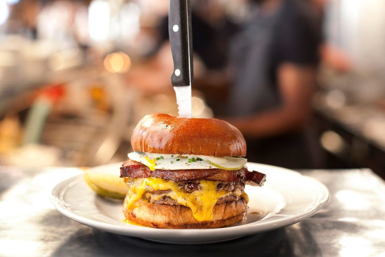 Burger impaled by knife on a plate, featuring bacon, cheese, and egg, in a bustling kitchen environment.