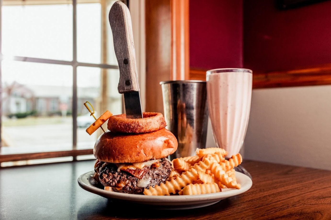 Burger with a knife stabbed through it sits on a plate of crinkle fries, next to a milkshake at a sunny diner table.