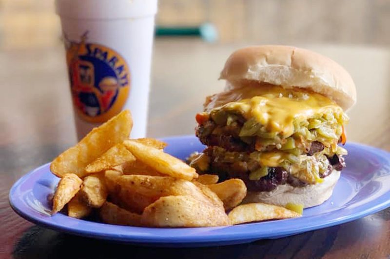 Burger topped with cheese sits beside crispy potato wedges on a blue plate, accompanied by a branded drink cup, on a wooden table.