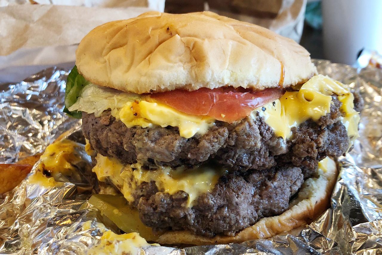 A double cheeseburger with lettuce and tomato sits on crinkled aluminum foil, surrounded by a blurred background.