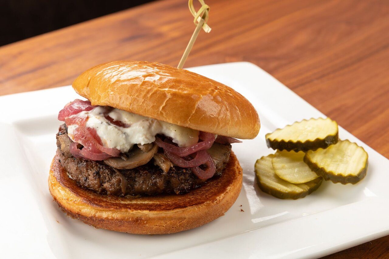 Burger sits on a white plate with sliced pickles, featuring cheese, sautÃ©ed onions, and mushrooms, secured by a skewer.