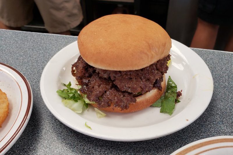 Burger sits on a white plate; layers of meat and lettuce within toasted bun; placed on a speckled gray countertop with nearby plate.