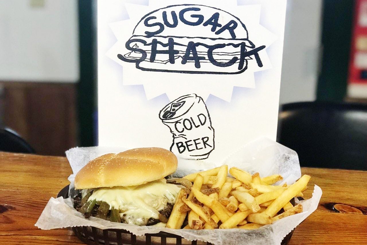 Burger with melted cheese and fries sit on a wooden table. "Sugar Shack" sign with a can reading "cold beer" in background.