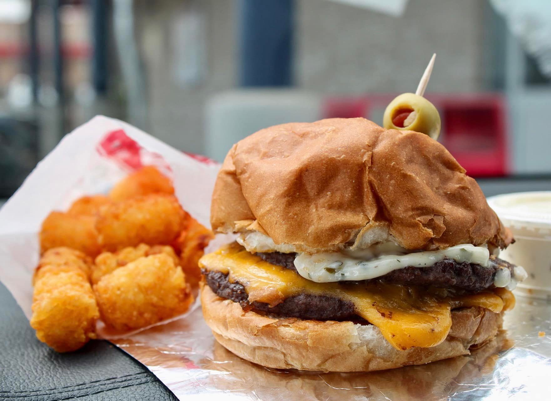 Cheeseburger sits on foil with a toothpick olive garnish; golden tater tots lie adjacent in open packaging on a table surface.
