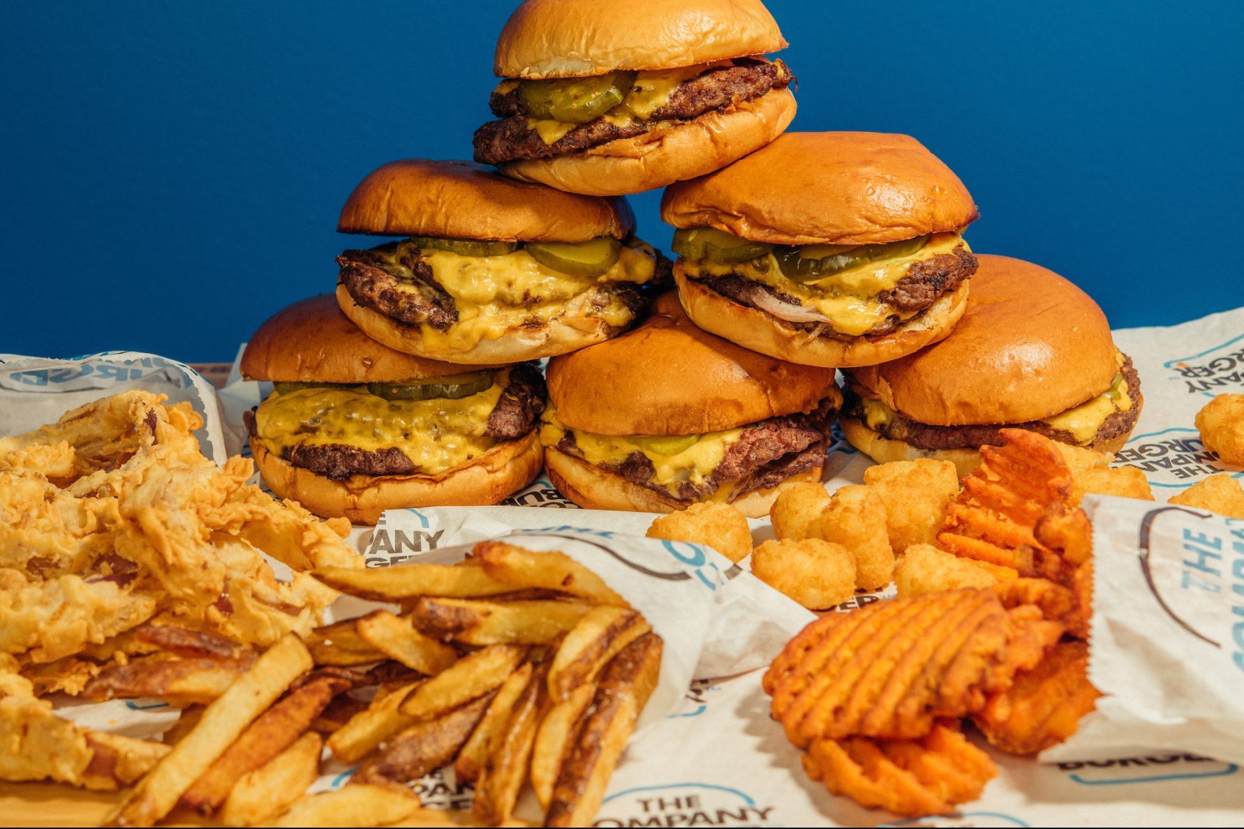 Burgers stacked with pickles and cheese sit on branded paper, surrounded by crispy fries, onion rings, and waffle fries against a blue background.