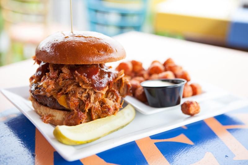 Burger overflows with shredded meat, displayed on a plate with tater tots, dipping sauce, and pickle, in a colorful, casual dining setting.