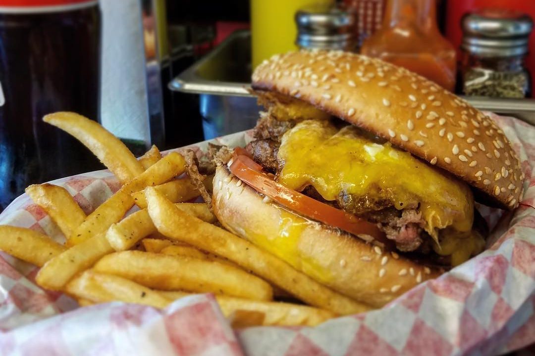 A cheeseburger with tomato sits on a checkered paper-lined basket beside golden fries, surrounded by condiments and a drink container.