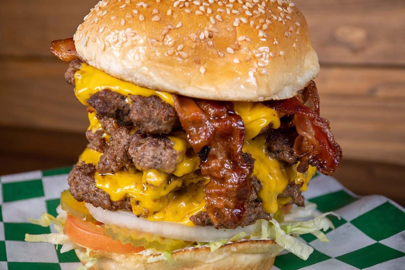 Burger stacked with beef, cheese, and bacon rests on lettuce and tomato, displayed on a green-checked paper in a wooden setting.