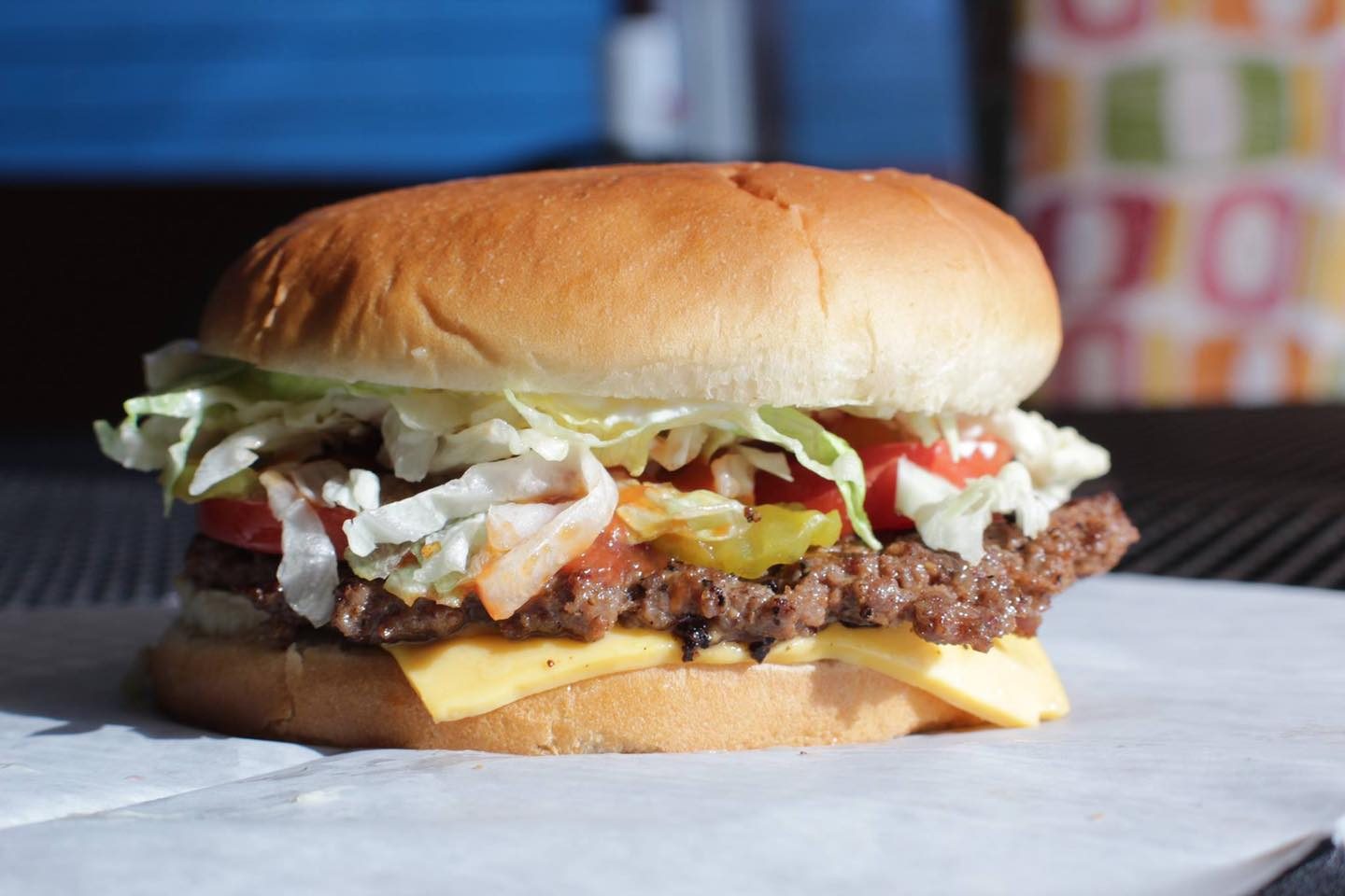Burger rests on parchment paper, featuring lettuce, tomato, cheese, and beef patty; background shows blurred colorful patterns and blue tones.