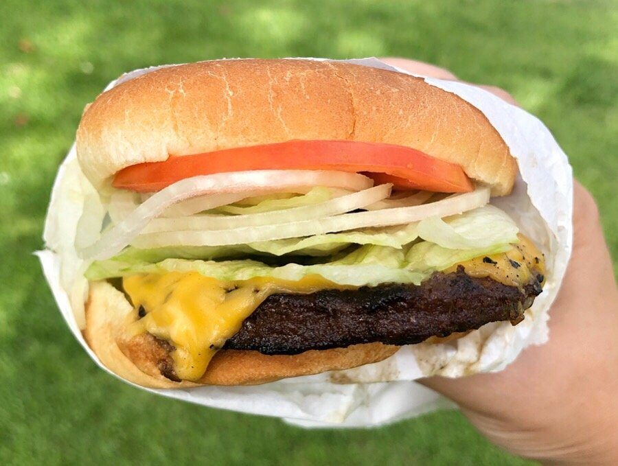 A hamburger with cheese, lettuce, tomato, and onion is held in hand, against a grassy background.