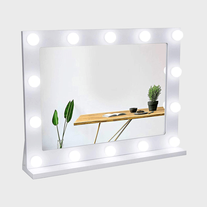 Mirror surrounded by bright bulbs reflecting a wooden table with plants and mugs against a plain white wall background.