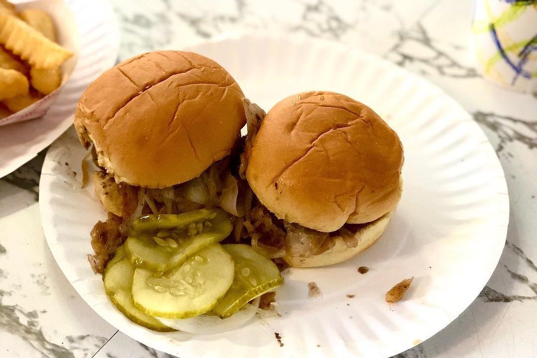 Two small burgers rest on a white paper plate with pickles, on a marble table. A side of crinkle fries is nearby.
