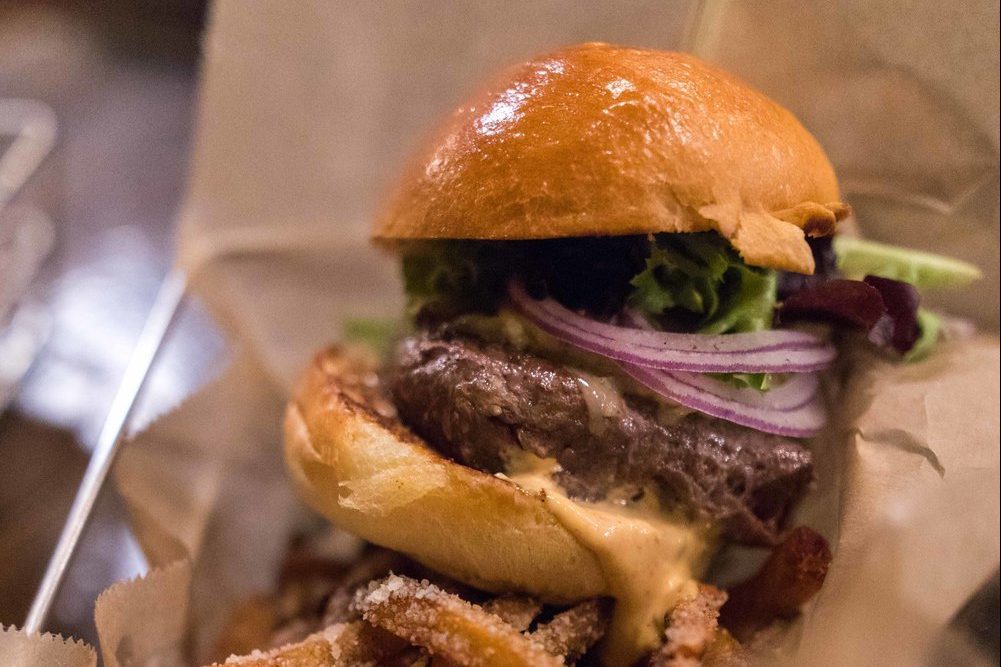 Burger rests on a paper wrapper, featuring a juicy patty, melted cheese, lettuce, and onions in soft bun, surrounded by fries.