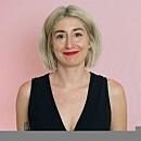 A person with short blonde hair smiles, wearing a black sleeveless top against a pink background.