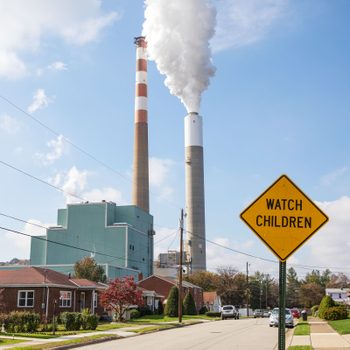 Two smokestacks emit clouds over residential houses with a "WATCH CHILDREN" sign on a quiet street. Bright day with clear blue sky.
