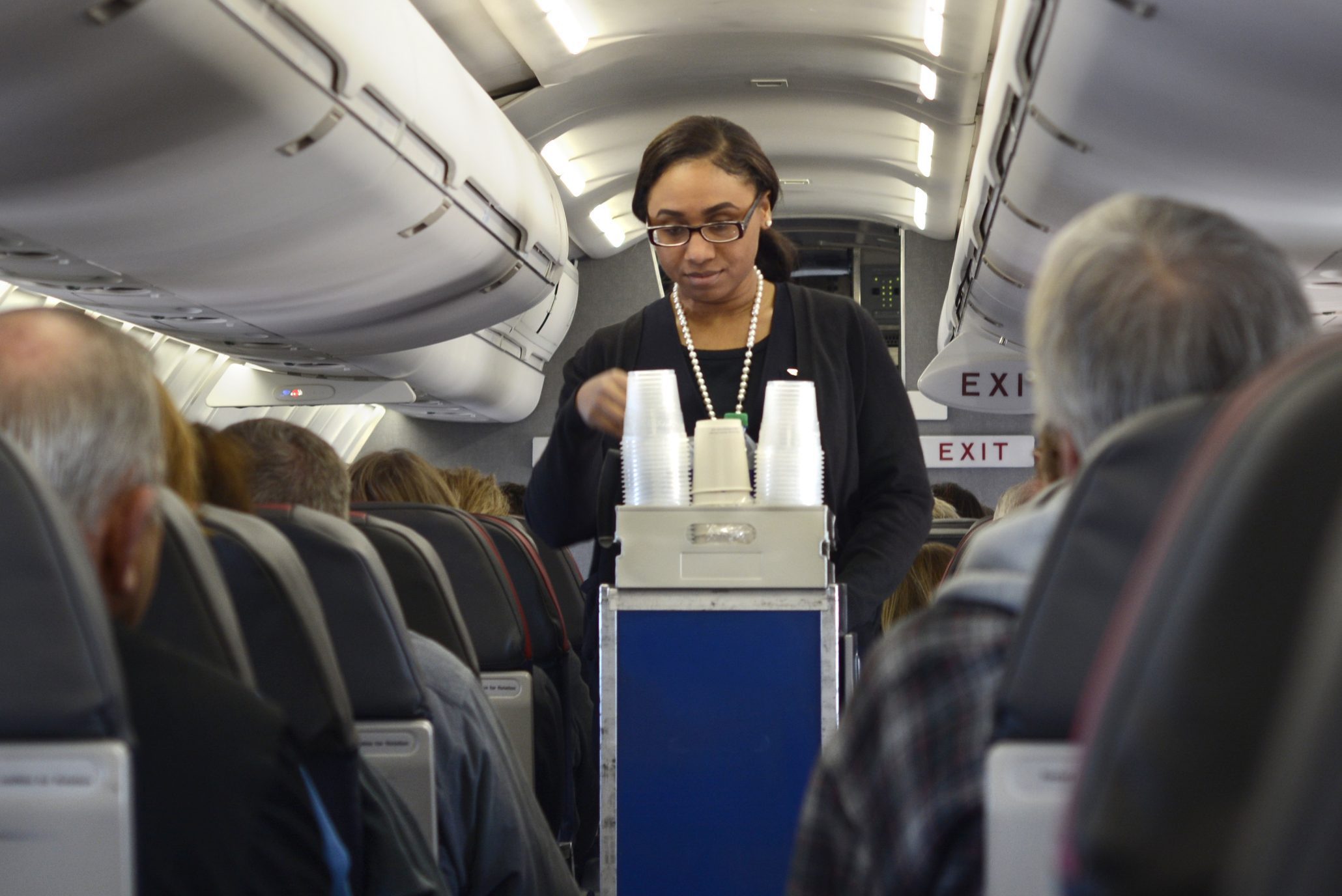 Flight attendant serves drinks from a cart in airplane aisle, surrounded by seated passengers, overhead bins, and exit signs.