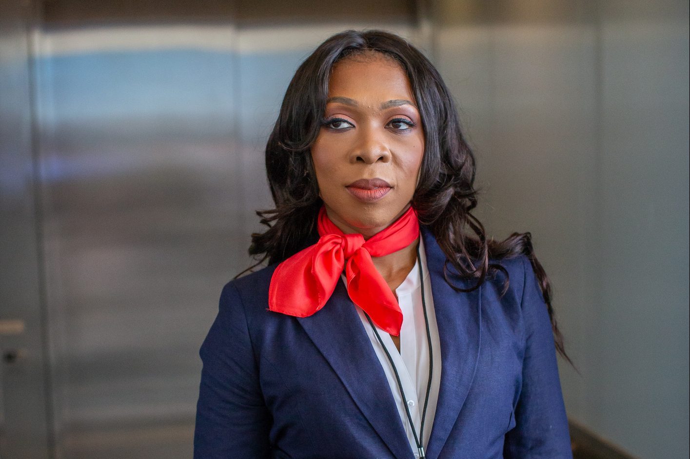 A woman stands, wearing a red scarf and blue blazer, in a modern indoor setting with blurred background.