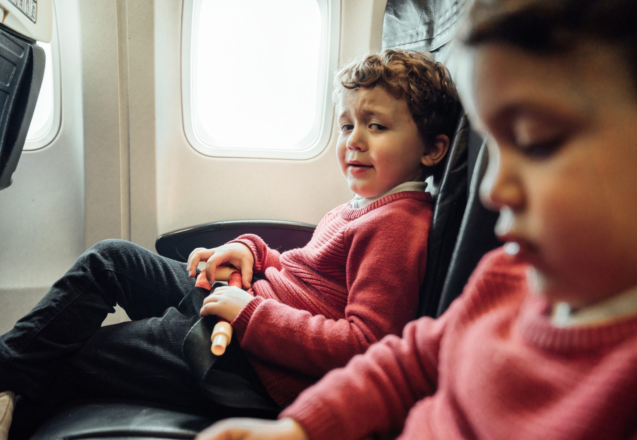 Child in red sweater holds a toy, sitting in an airplane seat next to another child, with a window visible behind them.