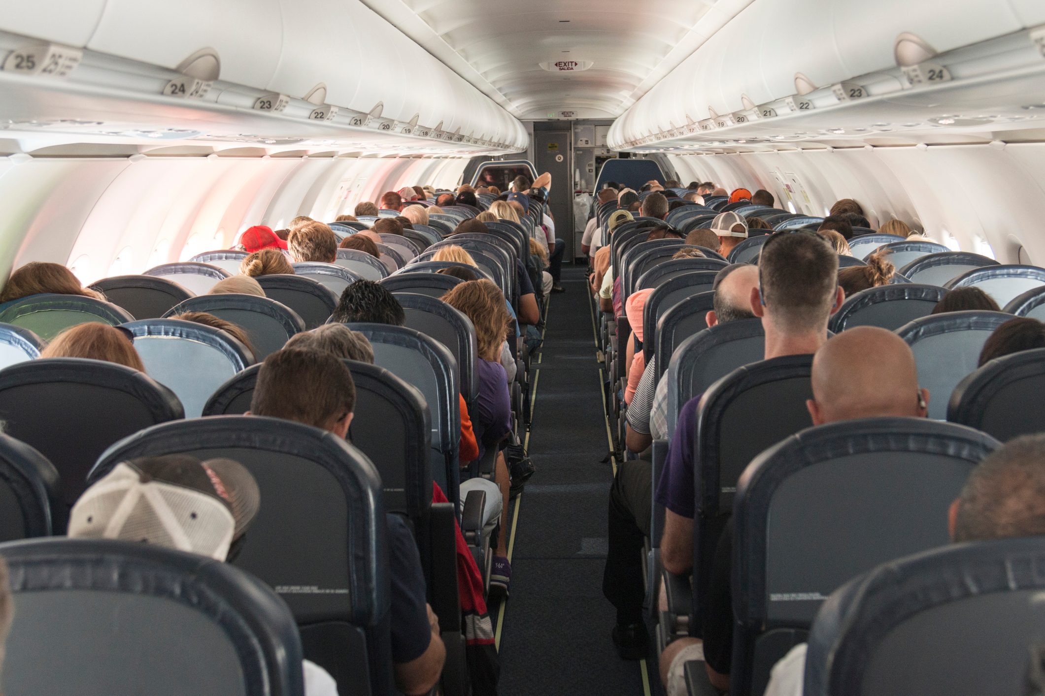 Passengers sit quietly in rows on an airplane, seated in a cabin with overhead compartments and an aisle running through the center.