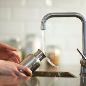 Hands fill a metal bottle with water from a kitchen faucet, in a tiled kitchen.