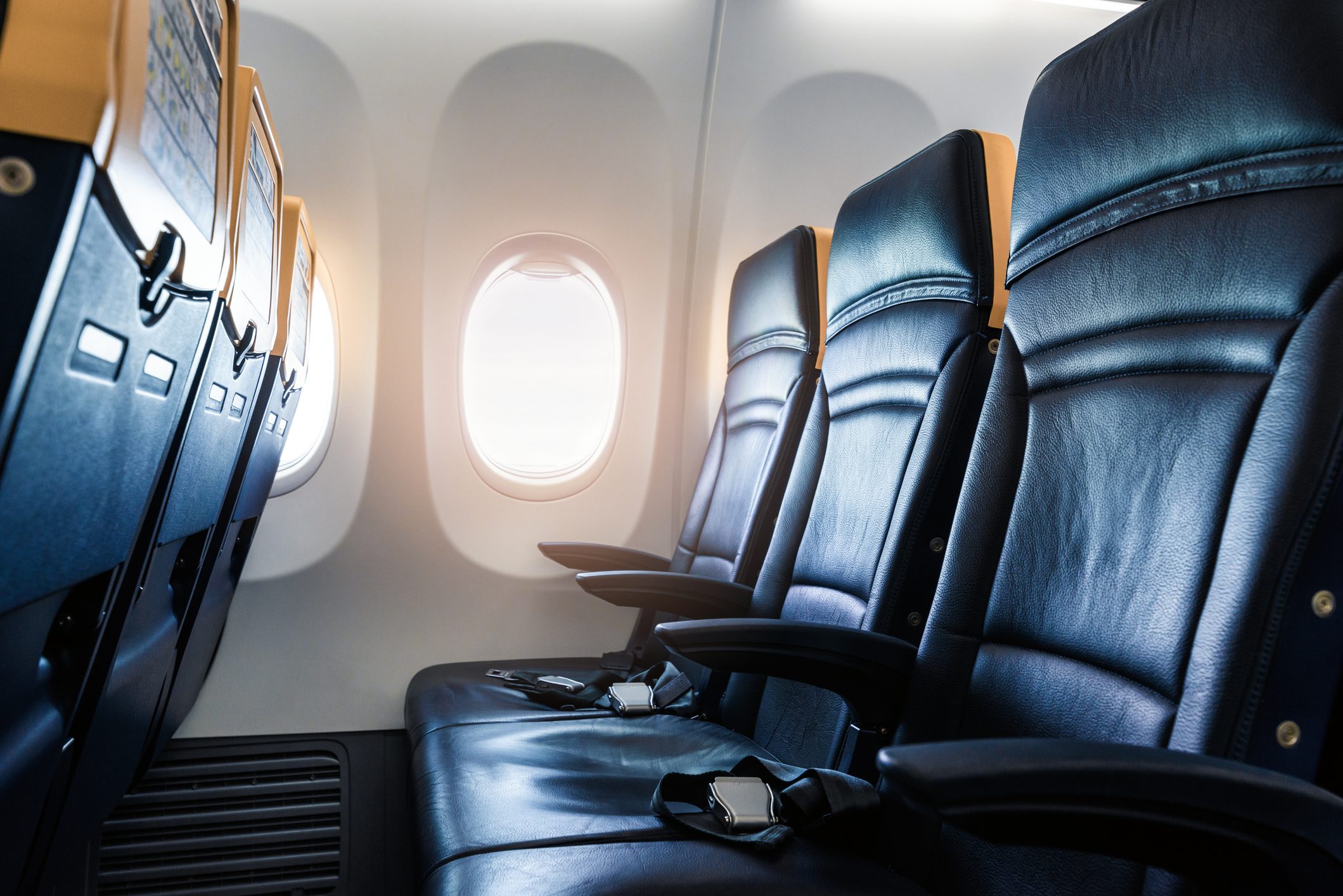 Airplane seats are aligned in rows, facing forward. Sunlight streams through oval windows, creating a bright cabin atmosphere. Seatbelts rest on blue cushions.