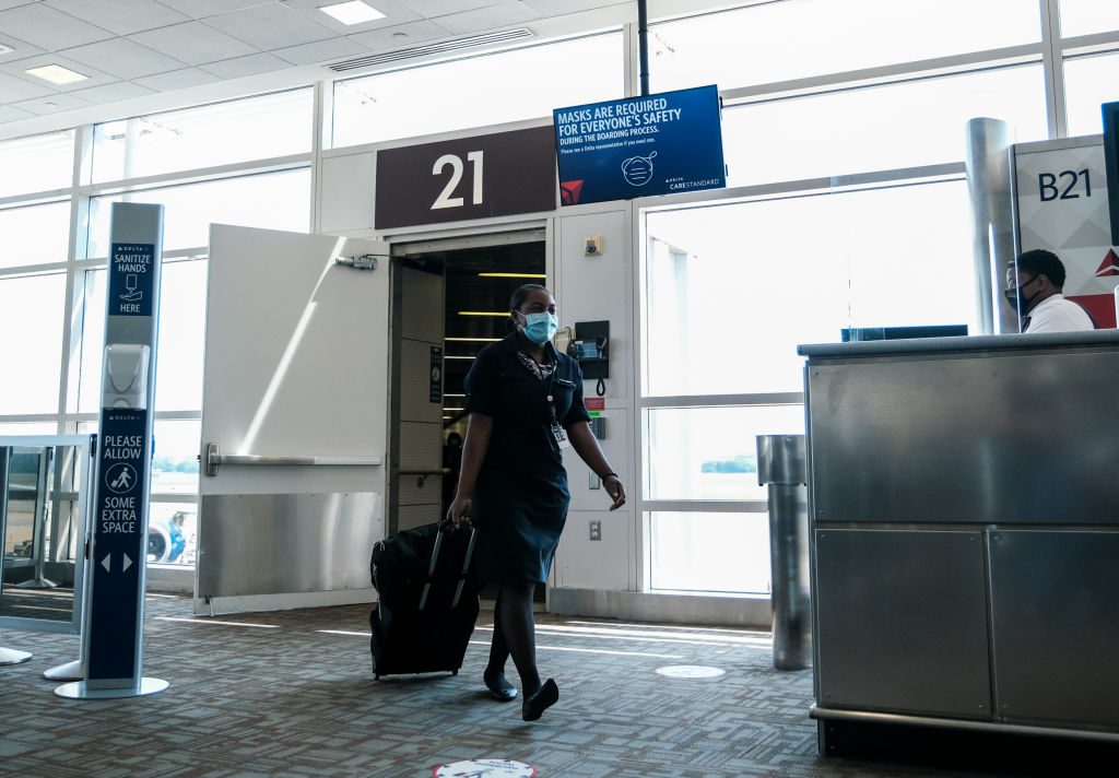 Person walks through airport gate holding luggage; wearing mask. Signs read "MASKS ARE REQUIRED," "PLEASE ALLOW SOME EXTRA SPACE," and "SANITIZE HANDS HERE."
