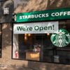 A Starbucks storefront displays a â€œWeâ€™re Open!â€ sign, with the logo visible. A poster encourages ordering ahead via the app.