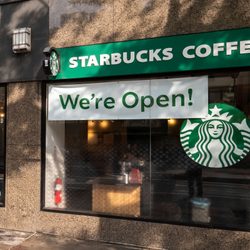 A Starbucks storefront displays a â€œWeâ€™re Open!â€ sign, with the logo visible. A poster encourages ordering ahead via the app.