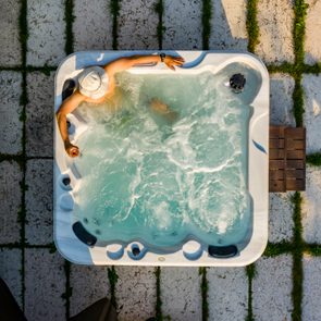 Person relaxes in bubbling jacuzzi, wearing a hat, holding a drink, surrounded by a grid-patterned stone patio and lush greenery.