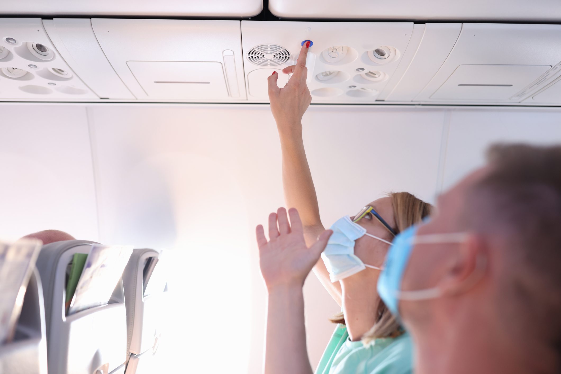 Passenger in a mask adjusts overhead controls on an airplane, reaching up from a seated position with another person nearby watching.