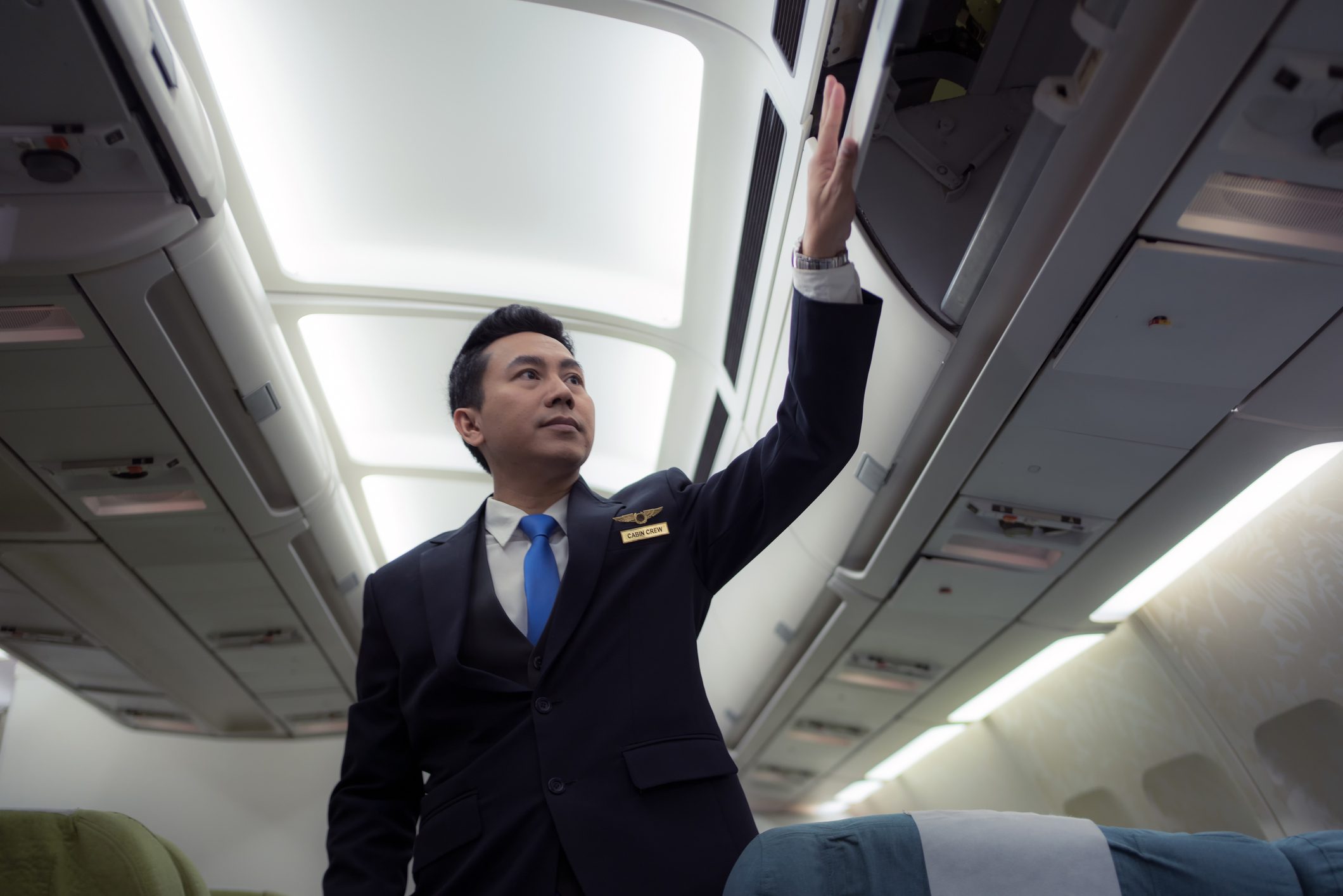 Cabin crew member, wearing a suit, closes overhead compartment in an airplane aisle, lit with bright ceiling lights.