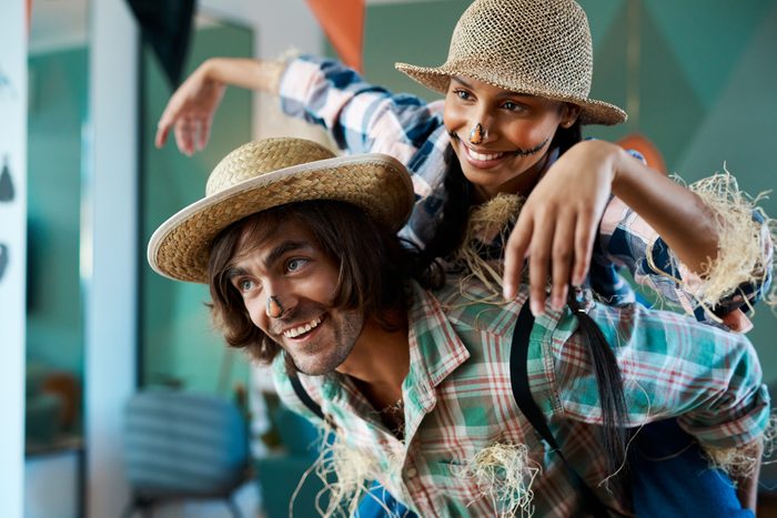 Two smiling people wearing straw hats and scarecrow costumes. One piggybacks the other in a brightly lit room. Native decorations are in the background.