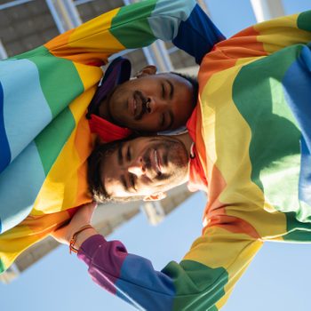 Two people embrace, wearing rainbow-colored clothing, against a clear blue sky with a building in the background.